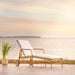 Natural rattan outdoor sun lounger with white cushions, rear wheels and a potted plant by the sea.