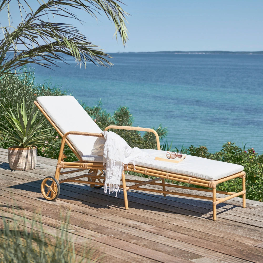 Natural rattan outdoor sun lounger with white cushions, rear wheels and adjustable backrest on a seaside deck.