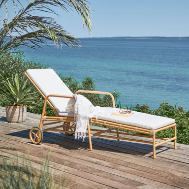 Natural rattan outdoor sun lounger with white cushions, rear wheels and adjustable backrest on a seaside deck.