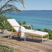 Natural rattan outdoor sun lounger with white cushions, rear wheels and adjustable backrest on a seaside deck.