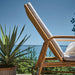 Natural rattan outdoor sun lounger with white cushions and rear wheels on a seaside patio.