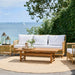 Natural teak outdoor coffee table with matching rattan sofa featuring white cushions on a seaside patio.
