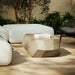 Gray geometric concrete outdoor coffee table, white sectional sofa and wicker chair with gray cushion on a tiled patio with greenery