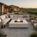 Outdoor patio furniture set in a vineyard landscape at sunset, featuring a large sectional sofa, matching armchairs, and a rectangular fire pit table with an active flame.