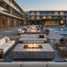 Modern outdoor patio furniture arrangement with concrete fire pits, white and beige sofas, and wooden armchairs around a swimming pool at dusk.