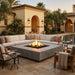 Outdoor patio scene with a large sectional sofa arranged around a square concrete fire pit table with visible flames. The setting includes patio pavers, a swimming pool, palm trees, and a stucco building with arches at dusk.