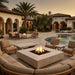 Outdoor patio scene at dusk with a large square fire pit centered in a curved beige cushioned sofa, surrounded by matching patio chairs and tables near a swimming pool and Spanish-style building with arches and palm trees.