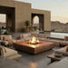 A modern outdoor patio scene with a large square fire pit table in the center, surrounded by low-profile sectional seating in light beige upholstery. The setting is a desert courtyard with concrete architecture, potted plants, and distant mountains at sunset.