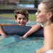 A smiling young man wearing a swim cap rests on a dark blue inflatable pool float in a swimming pool, with a woman in the foreground and a blurred outdoor background.