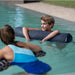 A boy wearing a swim cap floats on a dark gray cylindrical pool float in a swimming pool, with a woman in a blue swimsuit in the foreground.
