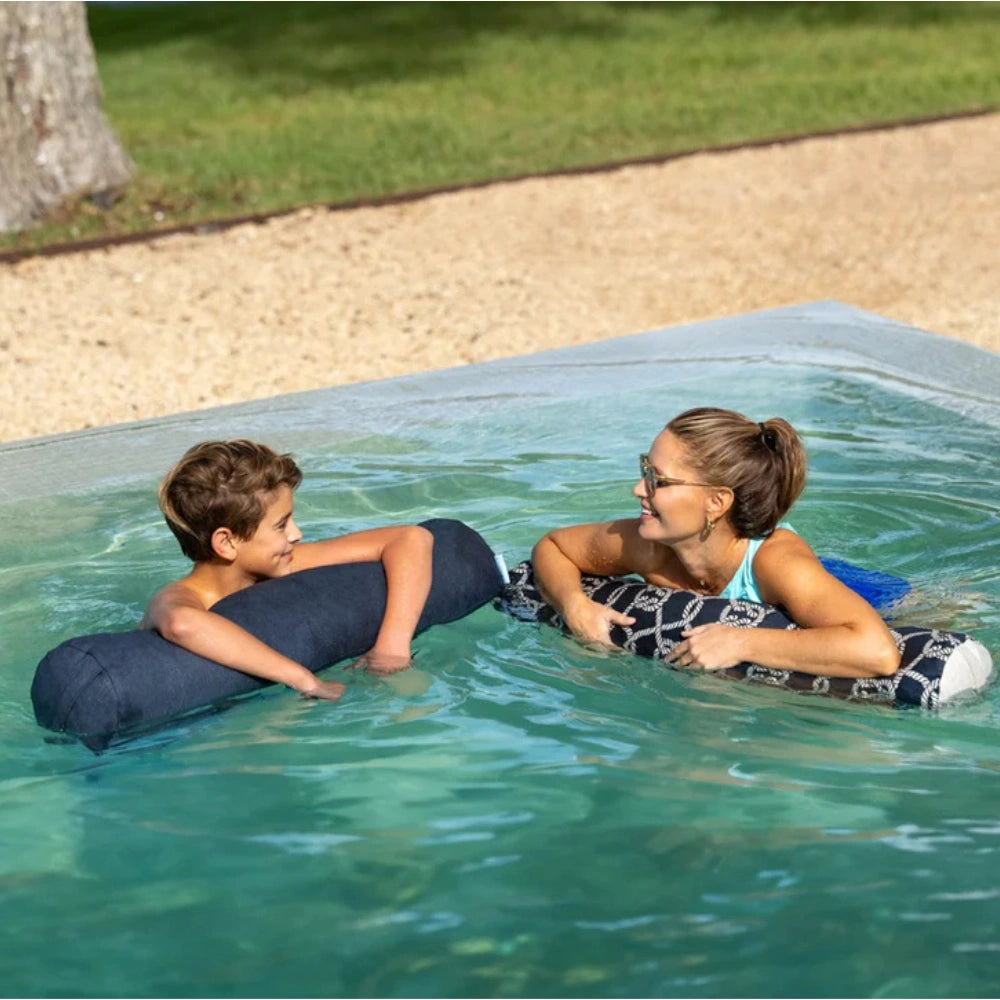 A woman and a boy relax in a swimming pool using inflatable pool floats. The woman wears a patterned black-and-white float and smiles at the boy, who is resting on a dark navy blue cylindrical float.