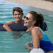 A woman in a blue swimsuit and sunglasses smiles in a swimming pool, with a young boy in the background also in the water.