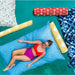A woman in a red bikini top and magenta bottoms relaxes on a large blue beanbag lounger. The scene is styled with colorful patterned pillows and rolled cushions on a bright teal surface.