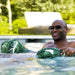 A smiling man wearing sunglasses relaxes in a swimming pool, holding two green and white tropical leaf-patterned float rings.