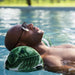 A man relaxing in a swimming pool, wearing a green and white tropical leaf-patterned float pillow and dark sunglasses.