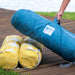 A person's hand is lifting a large teal-blue cylindrical carrying bag containing a yellow-and-white patterned cushion, set on a wooden deck outdoors.