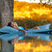 A woman relaxes on a blue inflatable lounge chair by a swimming pool at sunset, with a tree trunk and warm golden background visible.