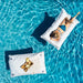 Two white rectangular pool float pillows floating in bright blue swimming pool water. One float has a woman reclining on it, and the other has a small food tray placed on it.