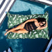 A woman in a black swimsuit relaxes on a large rectangular pool float with a green tropical leaf print, floating above a swimming pool.
