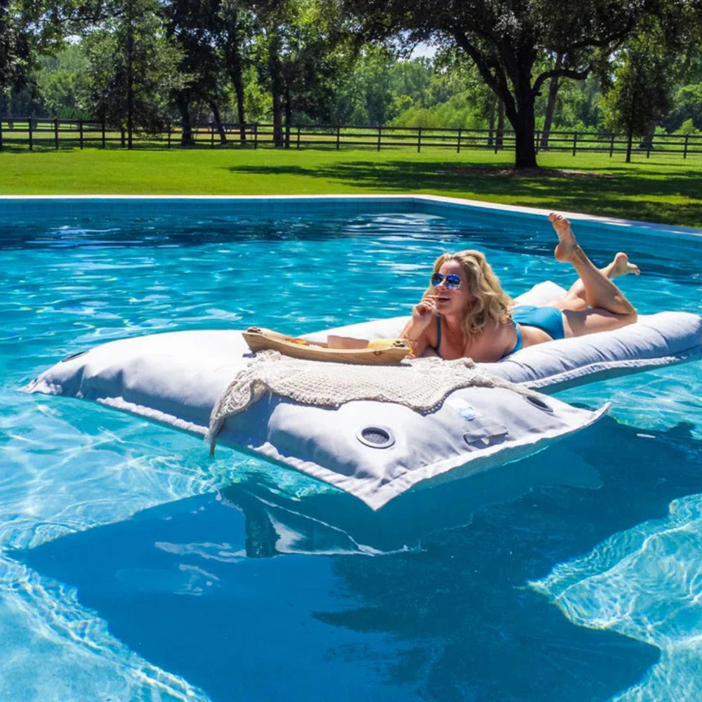 A woman wearing sunglasses relaxes on a large white inflatable pool float with a beige knit blanket, floating in a bright blue swimming pool. Trees and a grassy yard are visible in the background.