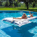 A woman wearing sunglasses relaxes on a large white inflatable pool float with a beige knit blanket, floating in a bright blue swimming pool. Trees and a grassy yard are visible in the background.