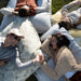 Four people lying on outdoor grass on oversized pillows and blankets, wearing knit cardigans and casual loungewear in neutral tones.