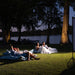 Outdoor evening scene on a grassy lawn by a lake, with several people relaxing under blankets and blue bean bags near large trees. A white screen on a stand is visible on the right, suggesting a movie or event setup.