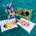 Four women lounging on oversized patterned pool float mats in a bright turquoise swimming pool, viewed from above.