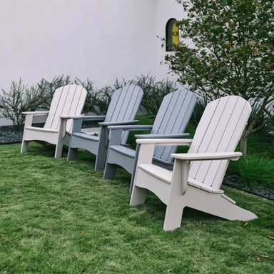 Four outdoor Adirondack chairs in white, light gray, and medium gray arranged on a green lawn in front of a white wall with shrubs and a tree.
