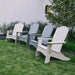 Four outdoor high density polyethylene resin Adirondack chairs in white, light gray, and medium gray arranged on a green lawn in front of a white wall with shrubs and a tree.
