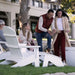 A lifestyle scene at an outdoor high density polyethylene resin patio showing a group of people relaxing in white Adirondack chairs. In the foreground, a man in a rust-colored puffer vest and blue jeans leans over a woman wearing a cream knit cardigan, white top, and burgundy pants.