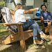 A woman sitting in a wooden textured high density polyethylene resin Adirondack chair pours coffee from a blue mug into a cup held by a man in another wooden chair, with an RV and outdoor grass in the background.