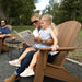 A woman and a young girl relax in a brown high density polyethylene resin Adirondack chair outdoors, with the woman reading a book and holding the child. Another person is seated at a small table in the background.