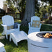 White outdoor patio furniture set including two Adirondack-style high density polyethylene resin chairs with matching ottomans and a round fire pit, arranged on a grassy lawn in front of a large tree.