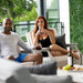A man and a woman relax in outdoor patio high density polyethylene resin chairs, holding wine glasses and a bottle of red wine on a table with food in the foreground. The setting features modern white lounge chairs, a green hedge wall, and large windows with green foliage outside.