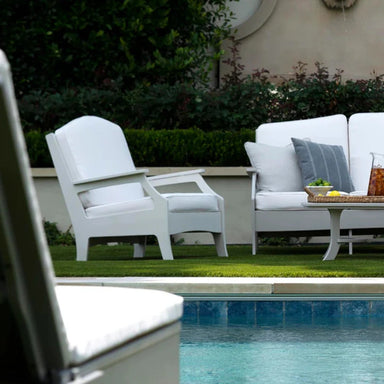 White outdoor club chair and sofa and a matching white sofa with gray and white pillows, set on a green lawn beside the pool.