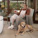 A woman relaxes in a light blue outdoor  high density polyethylene resin lounge chair on a patio, wearing a white apron over a rust-brown top and cream pants, holding a glass of red wine. A golden retriever lies on the tiled floor in front of the chair.