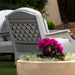 A light gray outdoor patio high density polyethylene resin chair with a lattice back panel, shown in a garden setting with a blurred planter of bright pink flowers in the foreground.