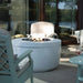 A round outdoor fire pit table with a white lid and visible fire rocks, set on a patio between high density polyethylene resin lounge chairs and a small side table with a glass of red wine.