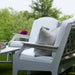 A white outdoor patio seating set with a high density polyethylene resin lounge chair, a matching armchair, and a side table on green grass, with a blurred garden background.