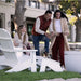 A family of three enjoys a sunny outdoor patio scene with white high density polyethylene resin lounge chairs. A man in a rust-colored puffer vest and blue jeans crouches beside a woman in a cream cardigan and white top, while a young girl sits on a white chair nearby.