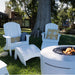White outdoor patio furniture set including two high density polyethylene resin Adirondack-style chairs, a matching ottoman, and a round fire pit table on a grassy backyard lawn with a tree and house in the background.
