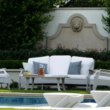 White outdoor patio high density polyethylene resin furniture set including a three-seat sofa, armchairs, and a coffee table, arranged on a lawn next to a swimming pool with a decorative stone lion wall in the background.
