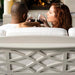 A couple sitting on a white outdoor high density polyethylene resin sofa, clinking wine glasses in front of a fireplace. The sofa features a decorative geometric lattice pattern on the front panel.