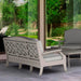 A light beige outdoor patio high density polyethylene resin loveseat with a decorative lattice backrest, shown next to a matching coffee table and gray lounge chair on a tiled patio with a garden view.