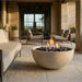 A round concrete fire pit bowl with a glowing flame and dark river rocks, placed on a stone patio in a modern outdoor living room setting.