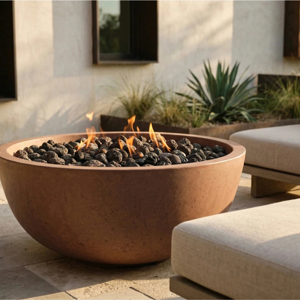 A large round terracotta fire pit with black lava rocks and visible flames, set outdoors on a stone patio beside light-colored lounge chairs and plants.