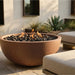 A large round terracotta fire pit with black lava rocks and visible flames, set outdoors on a stone patio beside light-colored lounge chairs and plants.