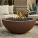 A large round terracotta-colored outdoor fire pit bowl filled with dark gravel and lit orange flames, set on a stone patio in a patio lounge setting.