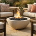 A round concrete fire pit bowl with black stones and a visible flame, placed on a stone patio between outdoor seating with neutral-toned cushions and a potted cactus in the background.